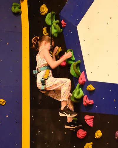 A young girl clipped into the auto-belay, climbing a black wall with green and pink holds