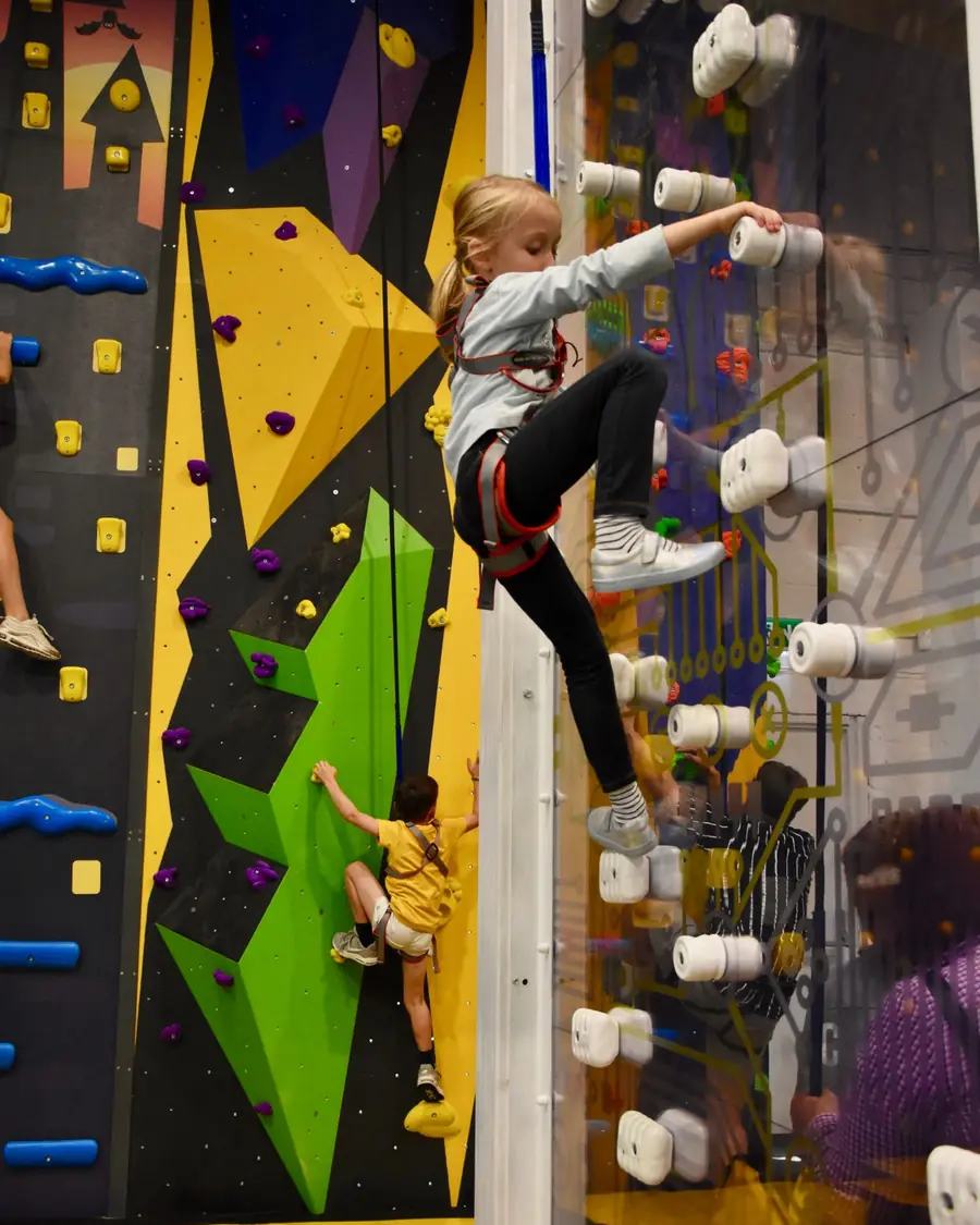 A girl in a harness climbing the see-through Fun Wall while a boy climbs the green wall behind her